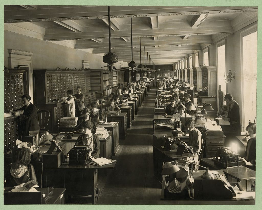 Photograph shows employees in the Library of Congress Copyright Office working at their desks and at the card catalog.